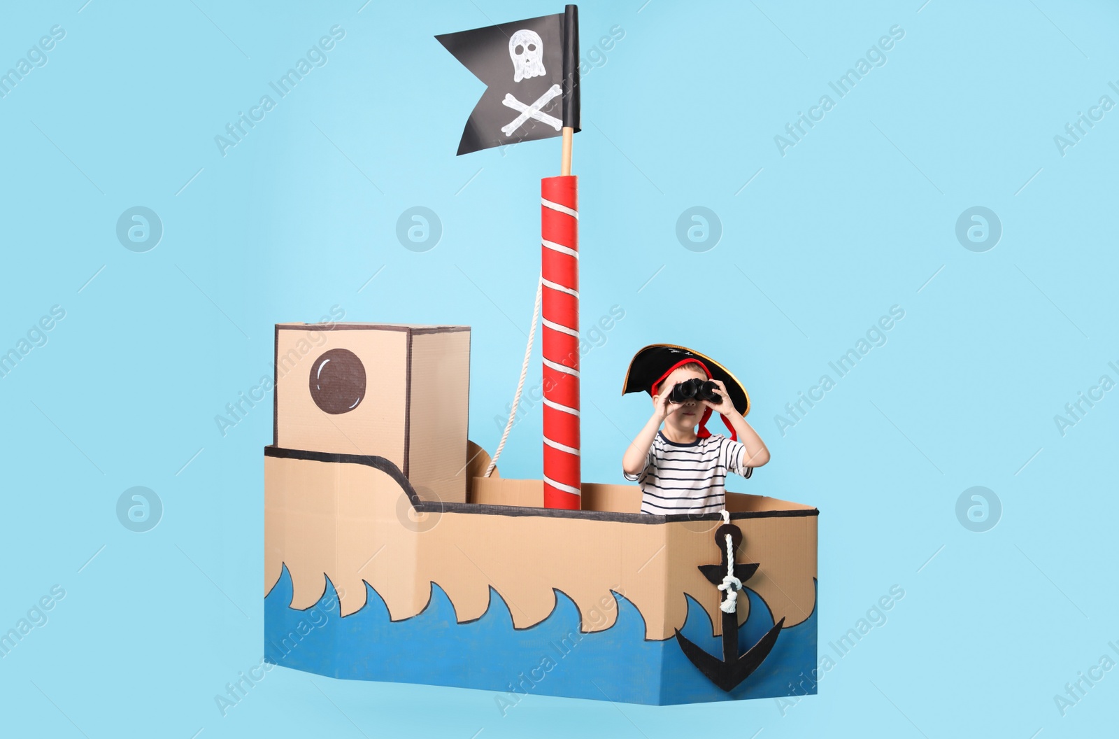 Photo of Little boy playing with binoculars in pirate cardboard ship on turquoise background