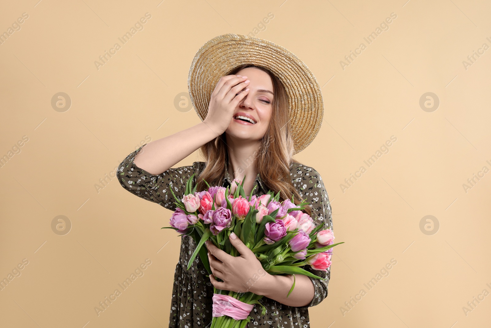 Photo of Happy young woman in straw hat holding bouquet of beautiful tulips on beige background