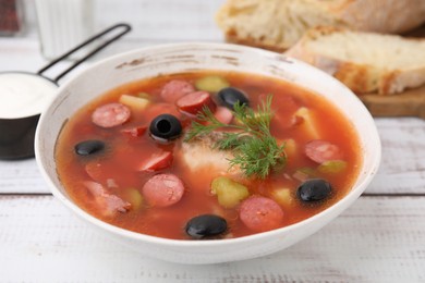 Photo of Meat solyanka soup with thin dry smoked sausages in bowl on white wooden table, closeup