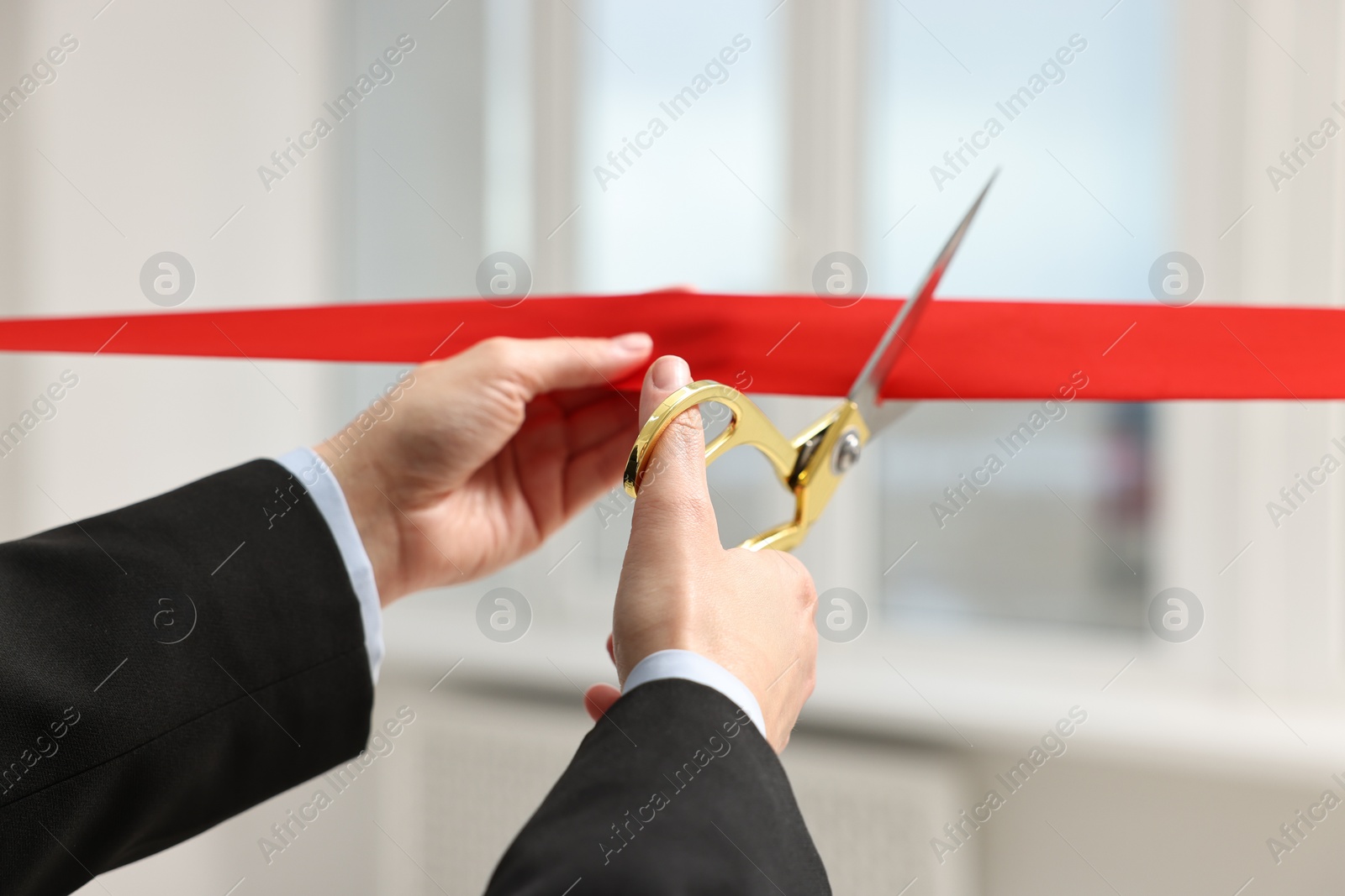 Photo of Woman cutting red ribbon with scissors indoors, closeup