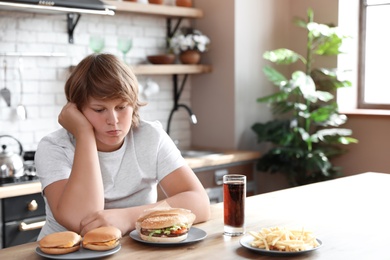 Emotional overweight boy at table with fast food in kitchen