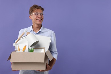Happy unemployed young man with box of personal office belongings on purple background. Space for text