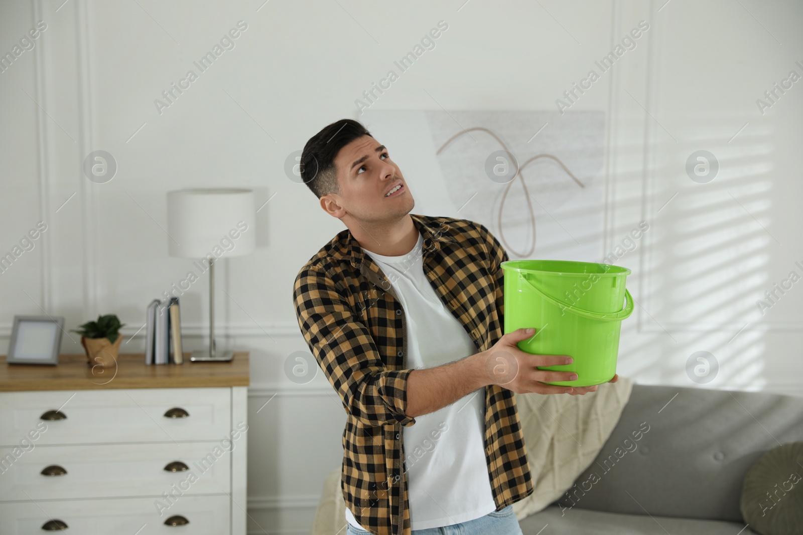 Photo of Emotional man collecting water leaking from ceiling in living room. Damaged roof