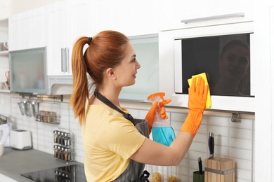 Photo of Woman cleaning microwave oven with rag and detergent in kitchen