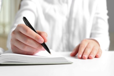 Photo of Woman writing in notebook at white table in office, closeup