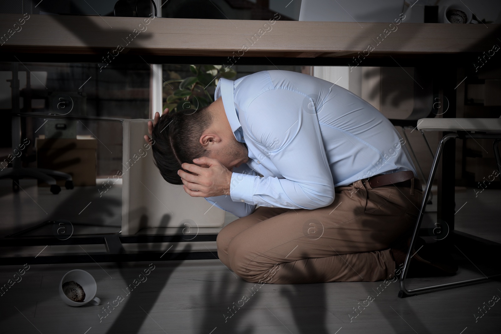 Image of Paranoid individual. Scared man hiding under desk and having delusion as hands reaching for him