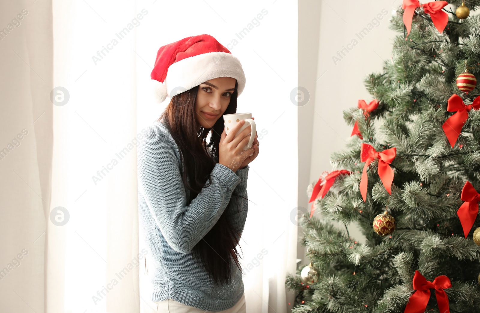 Photo of Beautiful young woman in Santa hat with cup of drink near Christmas tree at home