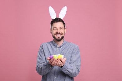 Happy man in bunny ears headband holding painted Easter eggs on pink background