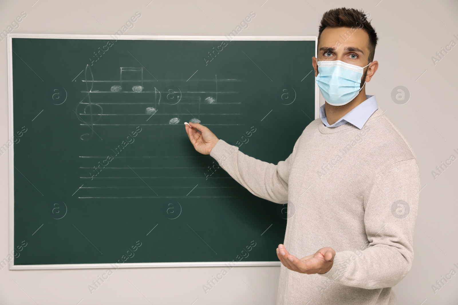 Photo of Teacher wearing protective mask near green chalkboard with music notes in classroom