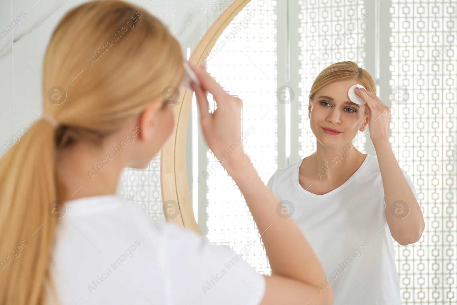 Photo of Happy young woman cleaning face with cotton pad near mirror in bathroom