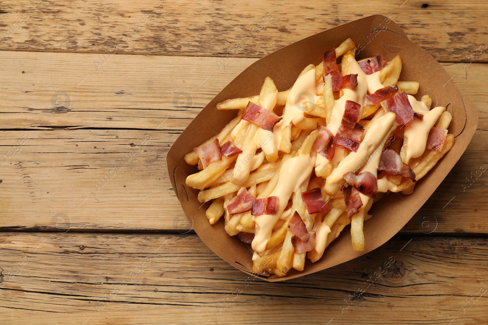 Photo of Tasty potato fries, cheese sauce and bacon in paper container on wooden table, top view. Space for text