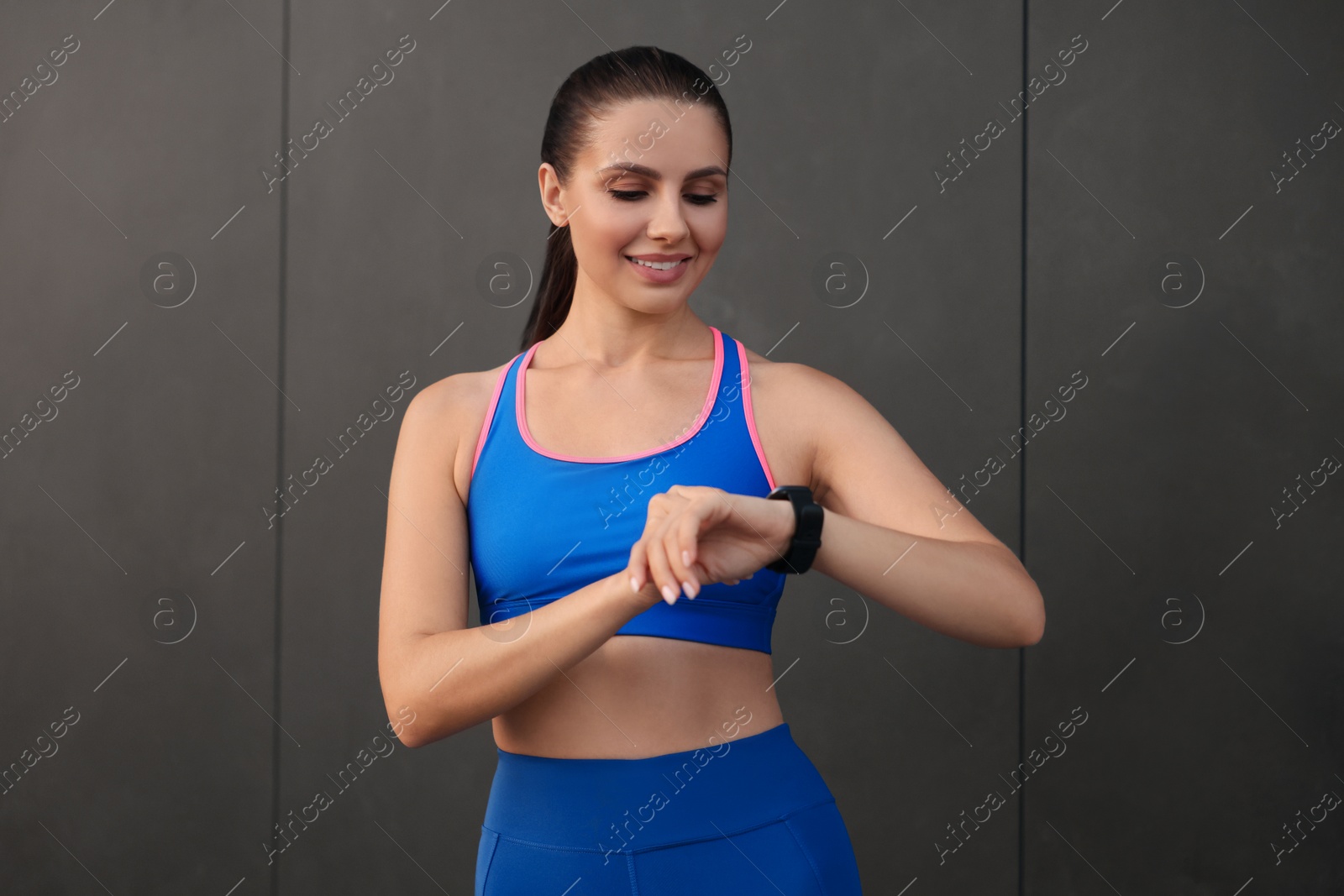 Photo of Beautiful woman in stylish sportswear checking smart watch near black wall