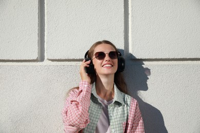 Photo of Smiling woman in headphones listening to music near white wall outdoors