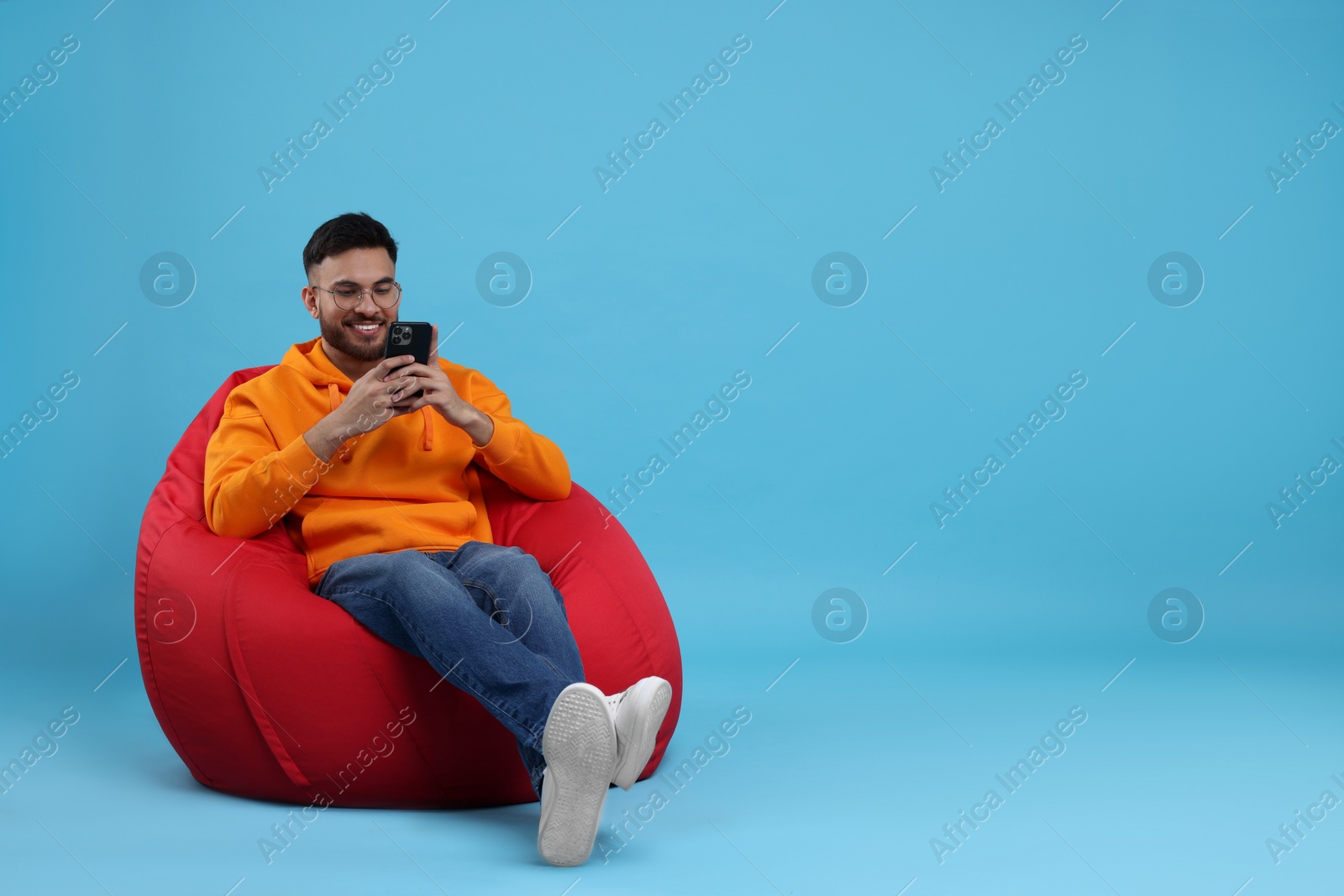 Photo of Happy young man using smartphone on bean bag chair against light blue background. Space for text