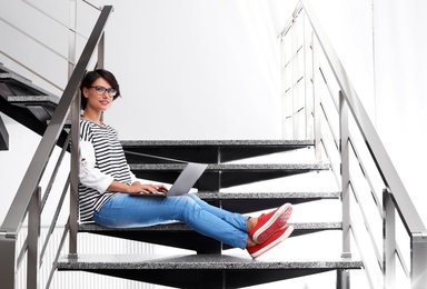 Young woman with modern laptop sitting on stairs indoors