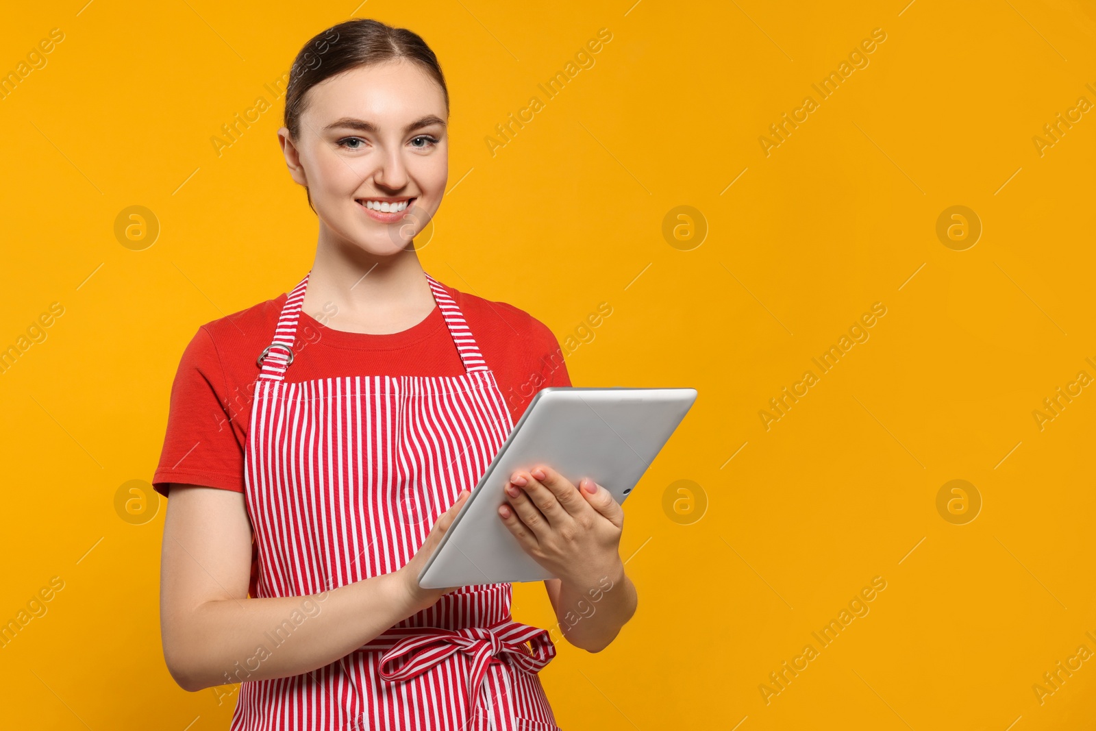 Photo of Beautiful young woman in clean striped apron with tablet on orange background. Space for text