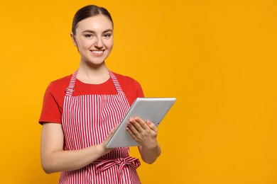 Beautiful young woman in clean striped apron with tablet on orange background. Space for text