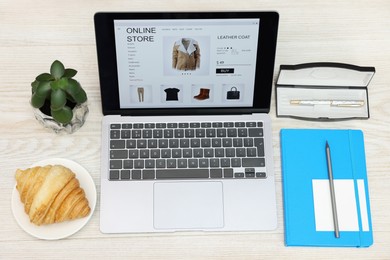 Online store. Laptop with open website, notebook, pen, croissant and pencil on wooden table, above view