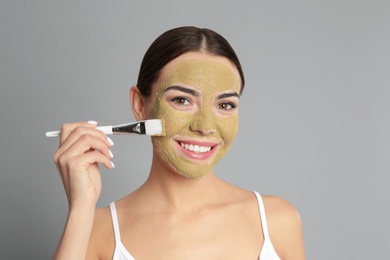 Photo of Young woman applying clay mask on her face against grey background