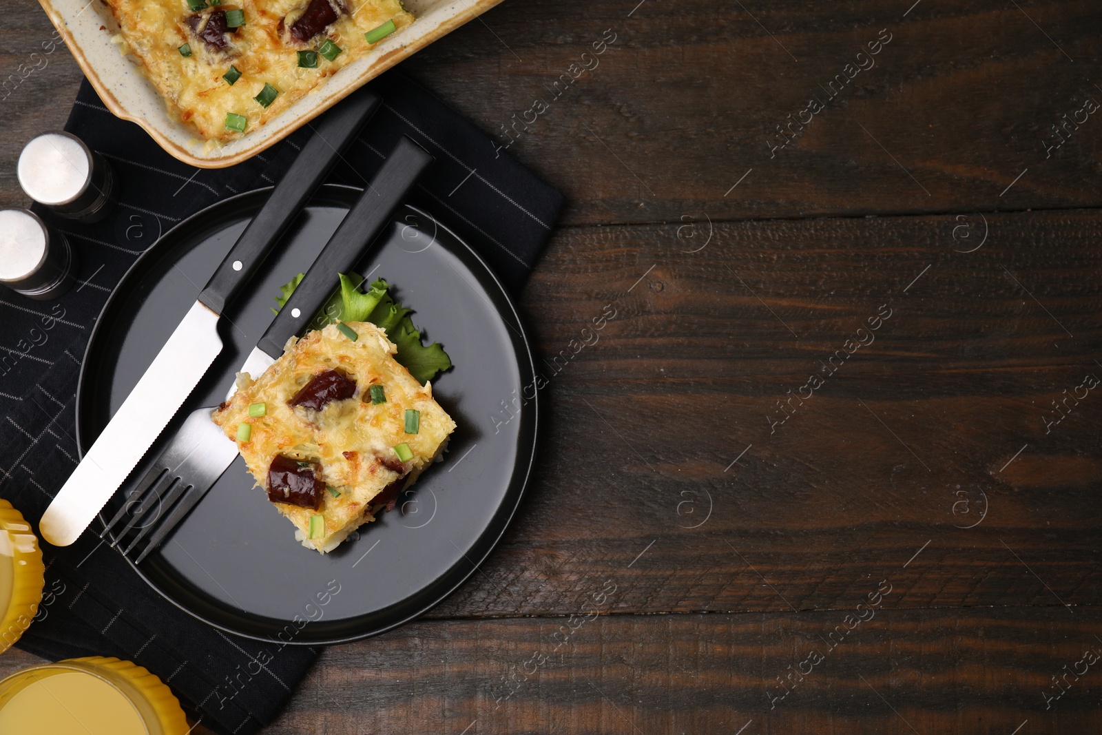 Photo of Tasty sausage casserole with green onions and cutlery served on wooden table, top view. Space for text