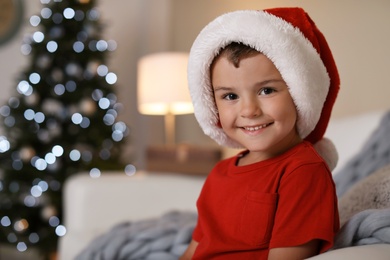 Photo of Cute little child in Santa hat sitting on sofa at home. Christmas celebration