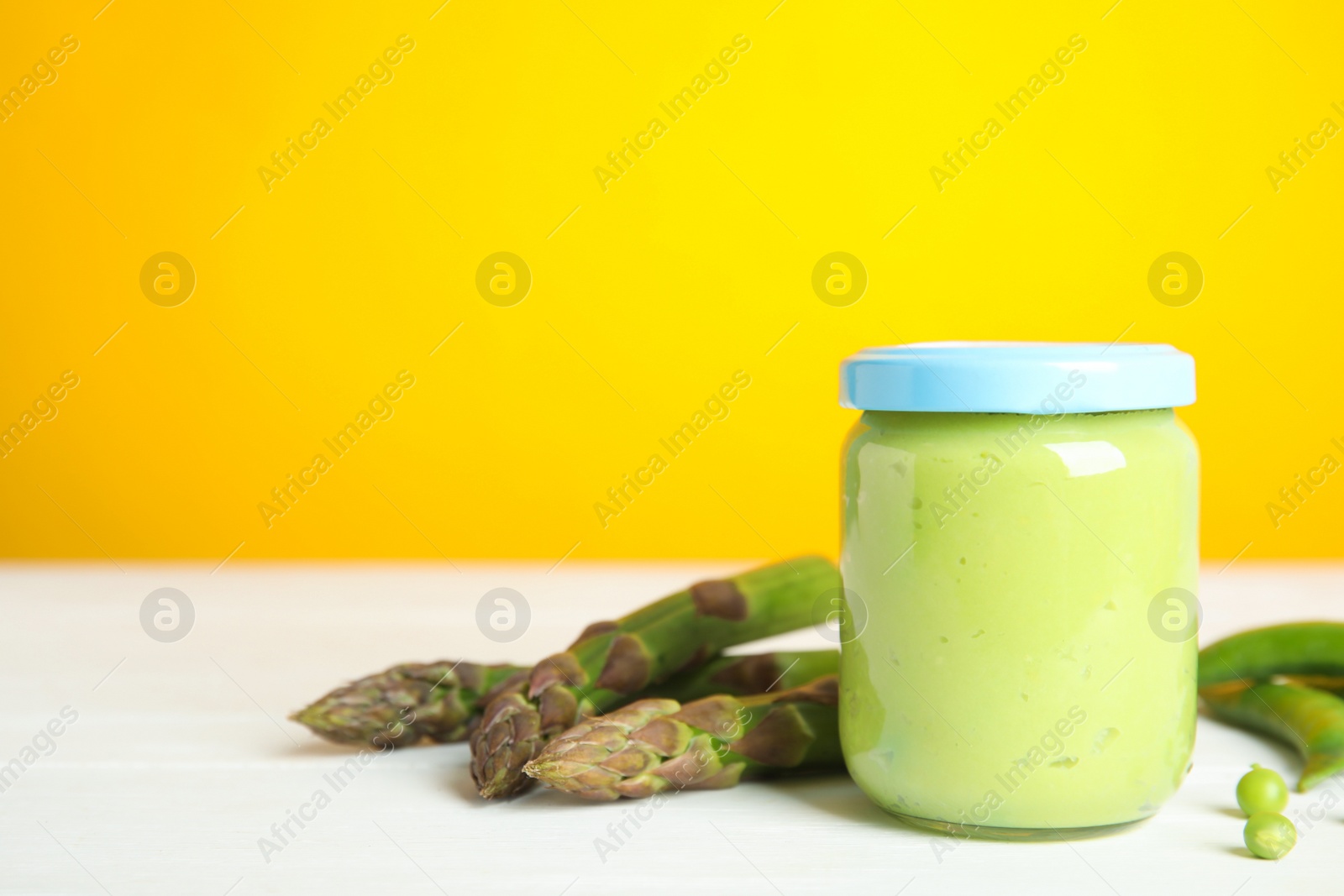 Photo of Jar with baby food, fresh pea pods and asparagus on white wooden table against yellow background, space for text