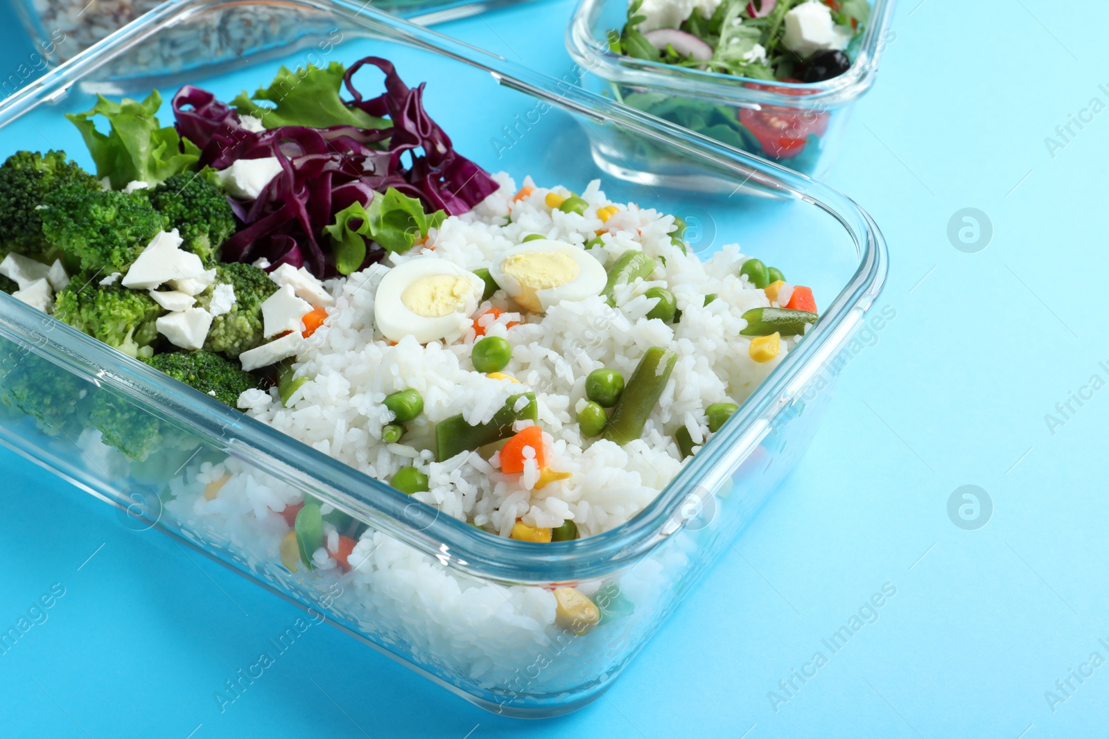 Photo of Tasty rice with vegetables and salad in glass container on light blue background