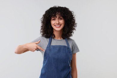 Photo of Happy woman pointing at kitchen apron on light grey background. Mockup for design