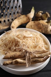 Photo of Plate with whole and grated horseradish roots on black wooden table, closeup