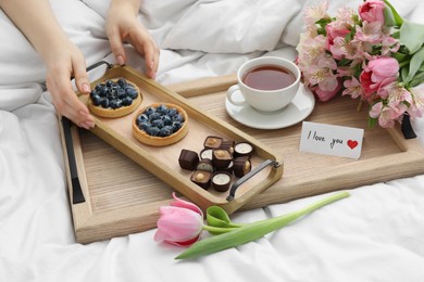 Photo of Tasty breakfast served in bed. Woman with desserts, tea, flowers and I Love You card at home, closeup