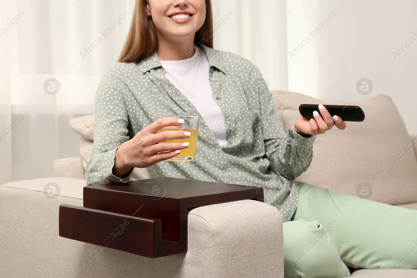 Photo of Woman holding glass of juice and remote control on sofa with wooden armrest table at home, closeup