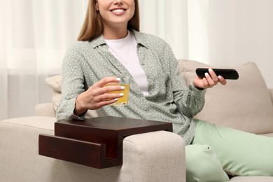 Photo of Woman holding glass of juice and remote control on sofa with wooden armrest table at home, closeup