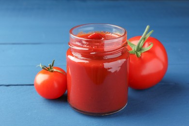 Photo of Jar of tasty ketchup and tomatoes on blue wooden table, closeup