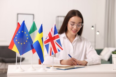Young woman writing in notebook at white table indoors, focus on different flags