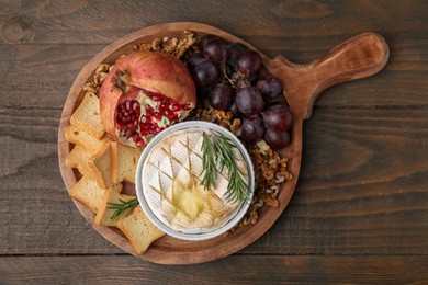 Photo of Board with tasty baked camembert, croutons, grapes, walnuts and pomegranate on wooden table, top view