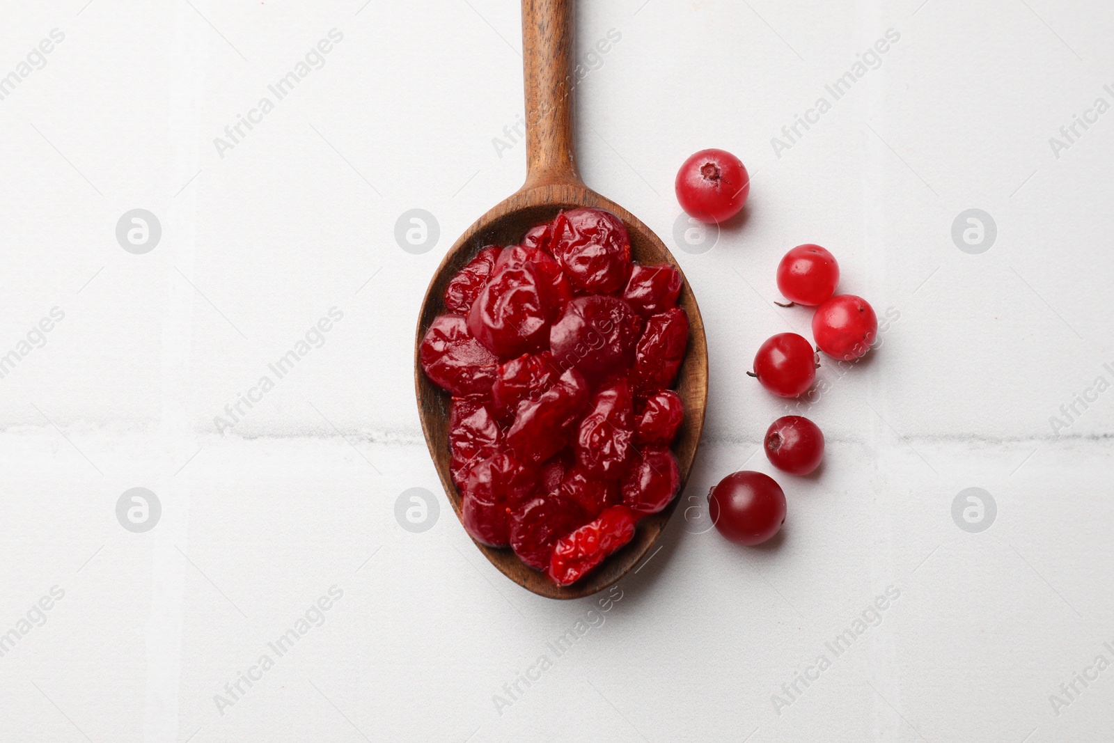 Photo of Tasty dried cranberries in spoon and fresh ones on white tiled table, top view