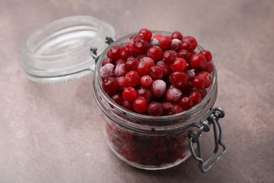 Photo of Frozen red cranberries in glass jar on brown textured table, closeup