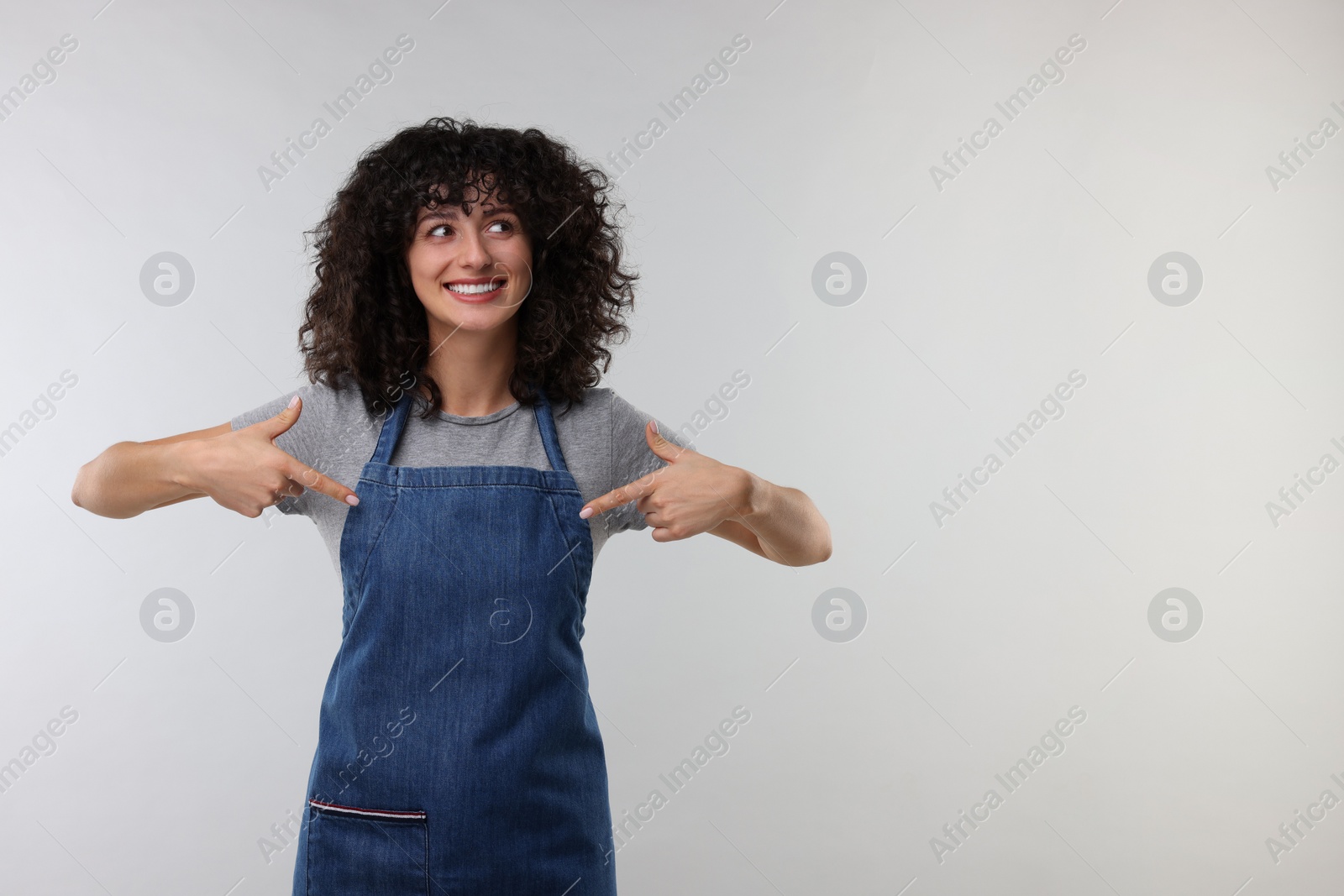 Photo of Happy woman pointing at kitchen apron on light grey background. Mockup for design
