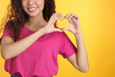 Photo of Happy African-American woman making heart with hands on yellow background, closeup