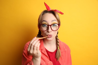 Photo of Fashionable young woman with braids and bright makeup chewing bubblegum on yellow background