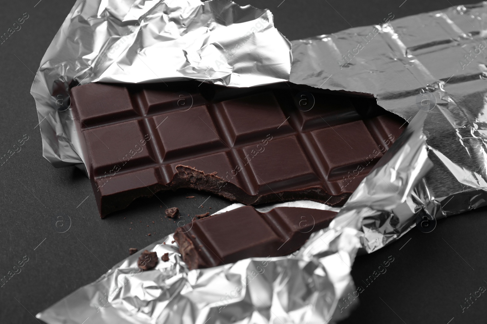 Photo of Broken dark chocolate bar wrapped in foil on black background, closeup