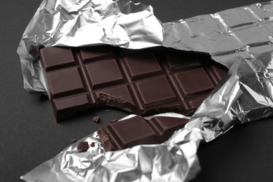 Photo of Broken dark chocolate bar wrapped in foil on black background, closeup