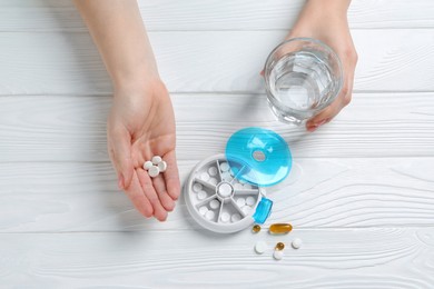 Woman holding glass of water and pills at white wooden table, top view