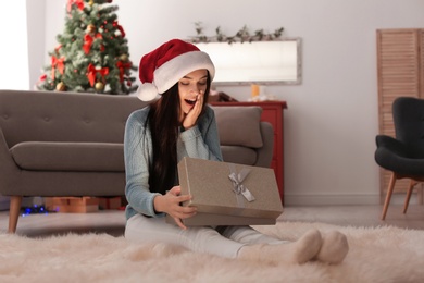 Photo of Beautiful young woman in Santa hat opening Christmas gift box at home