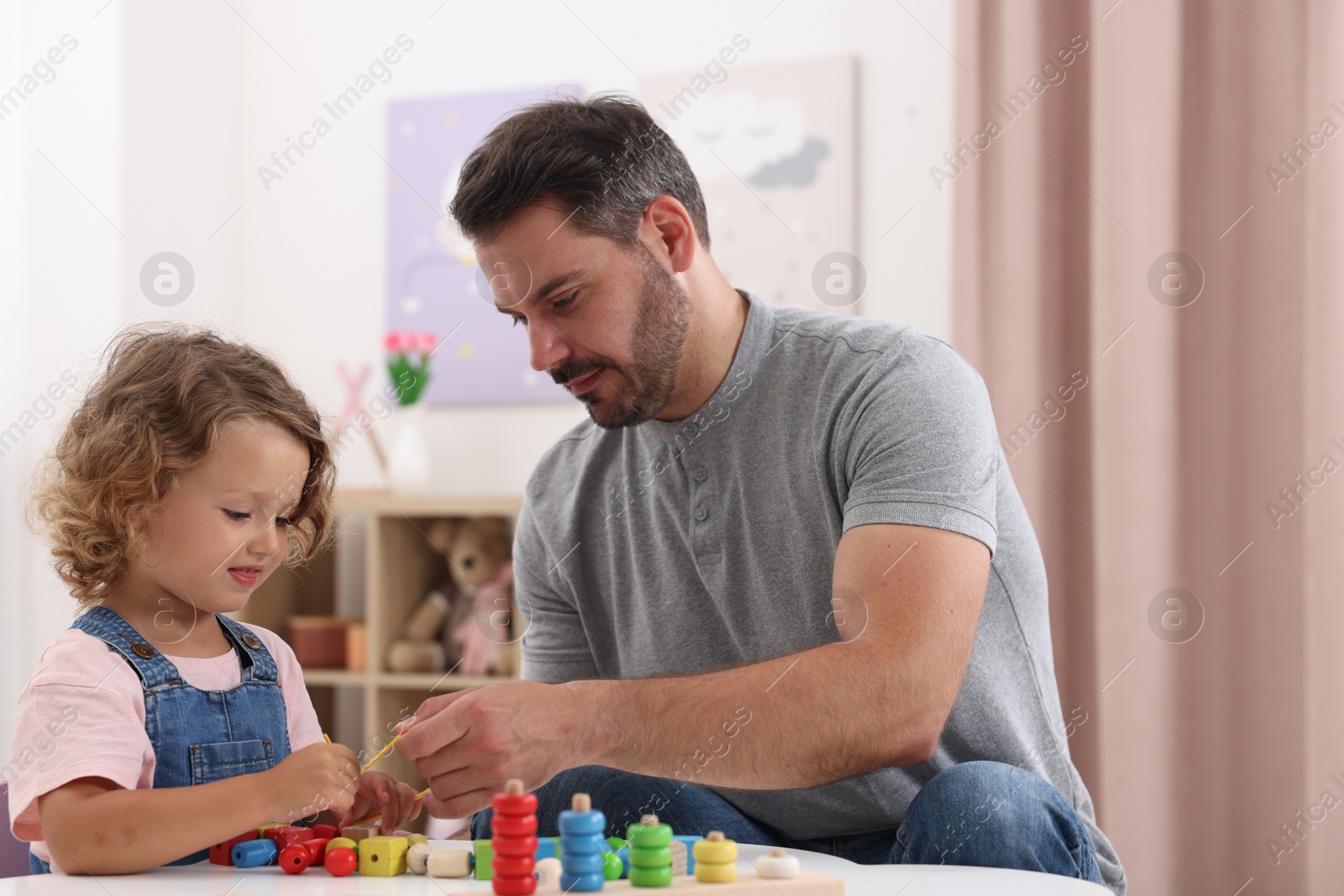 Photo of Motor skills development. Father and daughter playing with wooden pieces and string for threading activity at table indoors