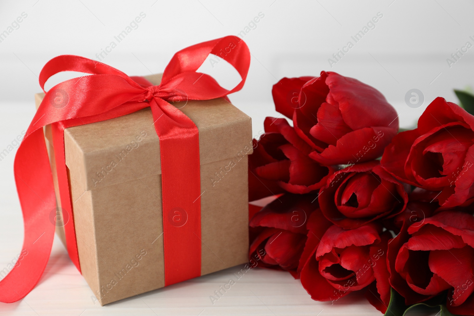 Photo of Beautiful gift box with bow and red tulip flowers on white table, closeup