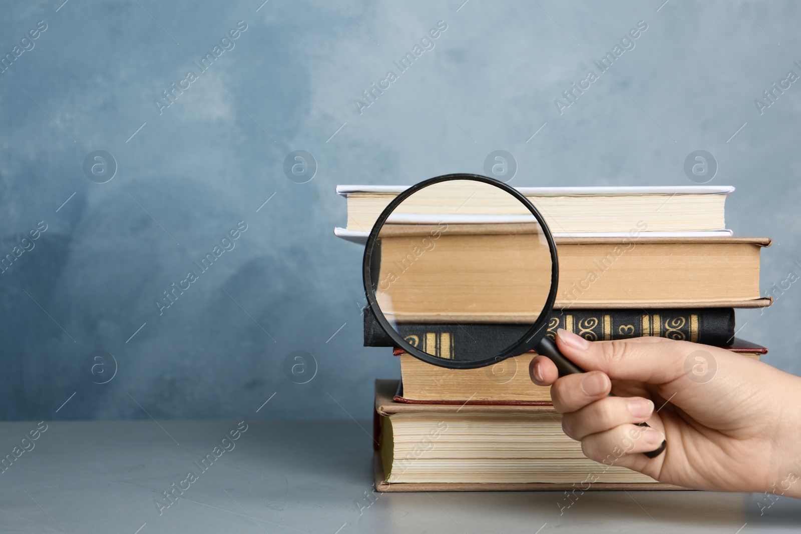 Photo of Woman looking through magnifying glass at stack of vintage books on grey table, closeup. Search concept