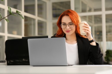 Photo of Happy woman with paper cup of coffee working on laptop at white desk in office