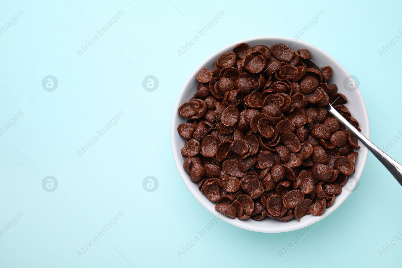 Photo of Breakfast cereal. Chocolate corn flakes in bowl and spoon on light blue table, top view. Space for text