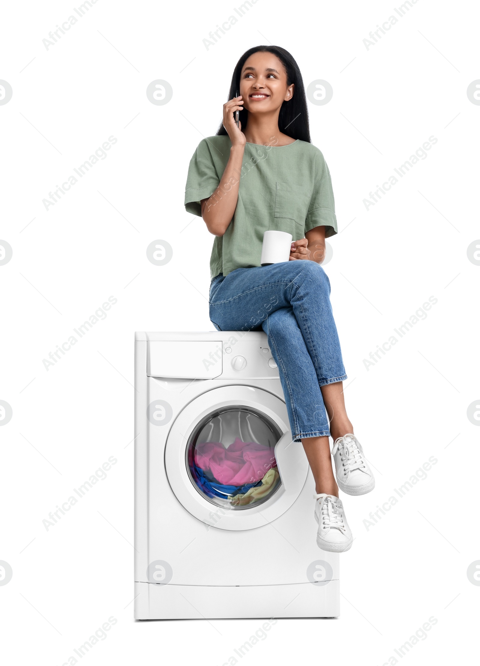 Photo of Beautiful woman with cup of drink sitting on washing machine and talking on phone against white background